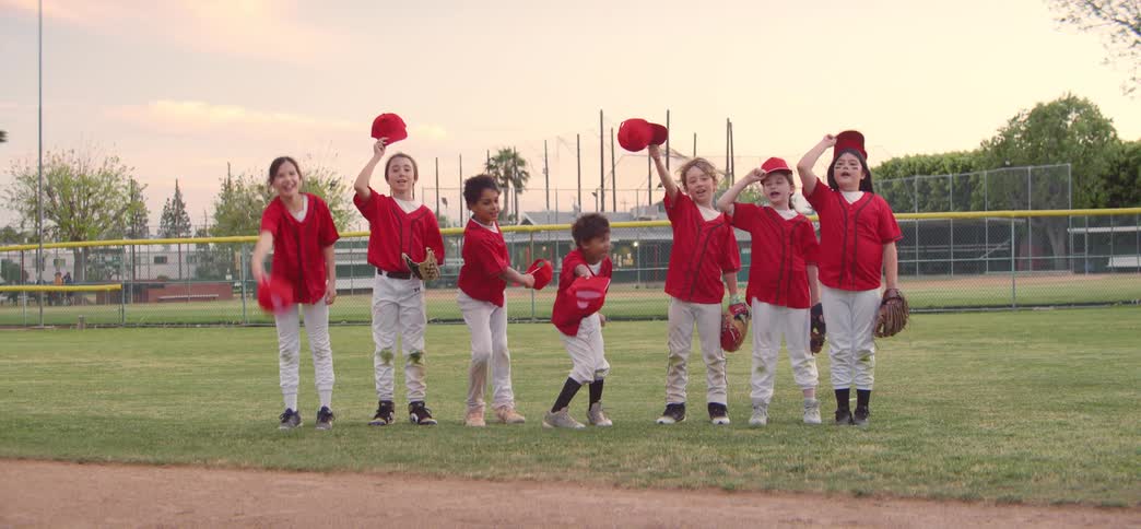 A diverse group of little league baseball players in red uniforms celebrate on the field by throwing their hats in the air in slow motion