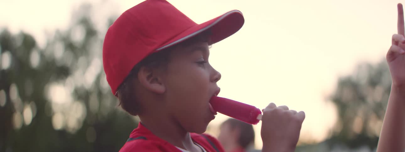 A young baseball player in a red uniform happily eats a popsicle with his teammate after a game in slow motion