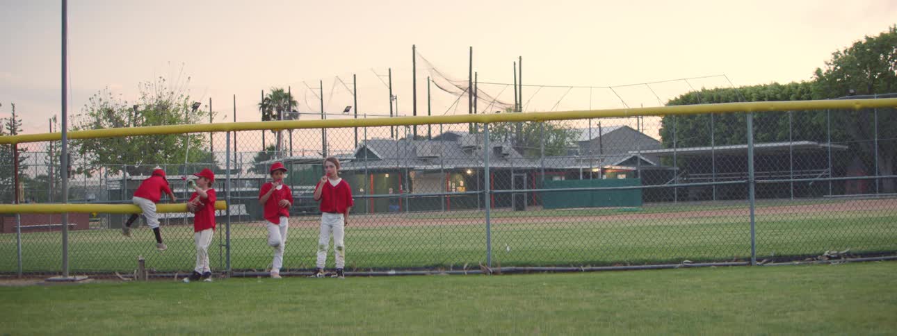 A group of young Little League baseball players in red uniforms watches the game from the outfield fence at dusk