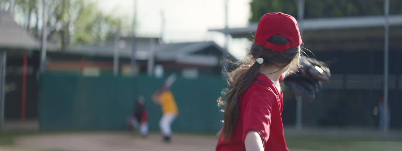 A young girl pitcher in a red uniform throws a baseball in slow motion, which the batter hits before running to first base