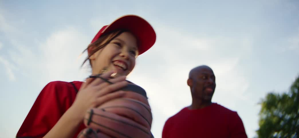 A happy young girl in a red uniform throws a baseball in slow motion as her father cheers her on from a low angle