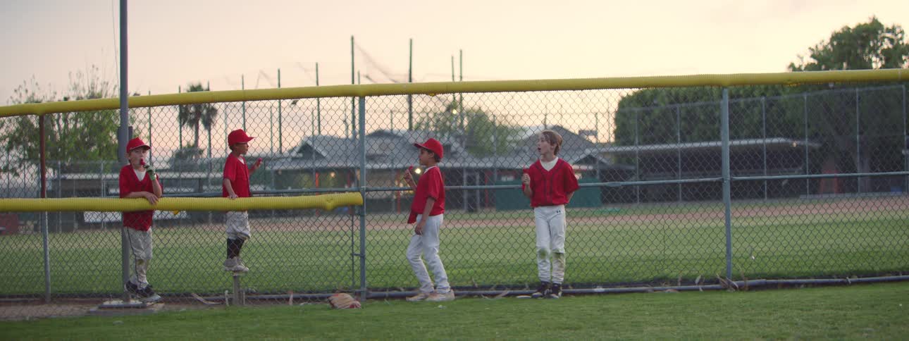 A group of young little league baseball players in red uniforms casually eat lollipops by the dugout fence at dusk