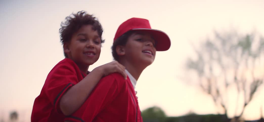 Two happy brothers in red baseball uniforms share a playful piggyback ride in slow motion at dusk