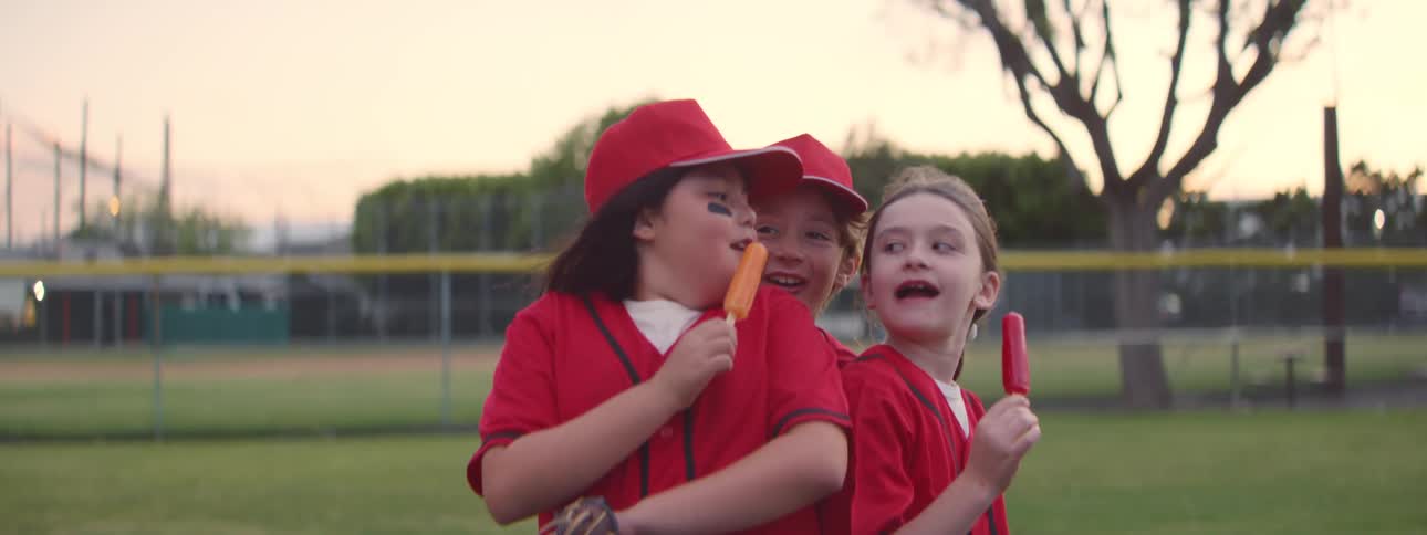 A group of happy little league teammates enjoy colorful popsicles together on the baseball field in slow motion