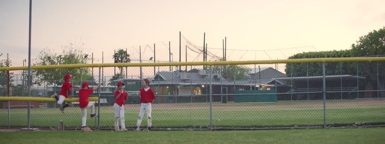 Four young boys in their red Little League uniforms wait by the chain-link fence of the baseball field as the sun sets