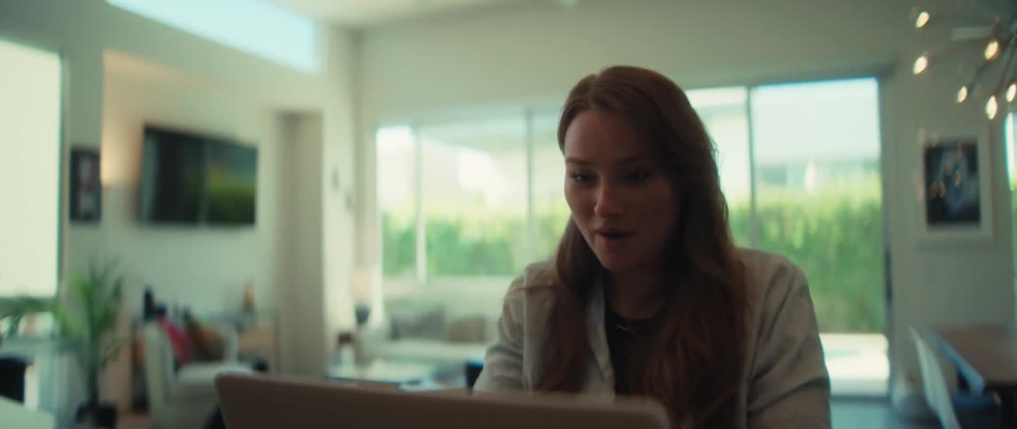 A smiling young woman on a video call reacts happily while working on her laptop from her bright living room