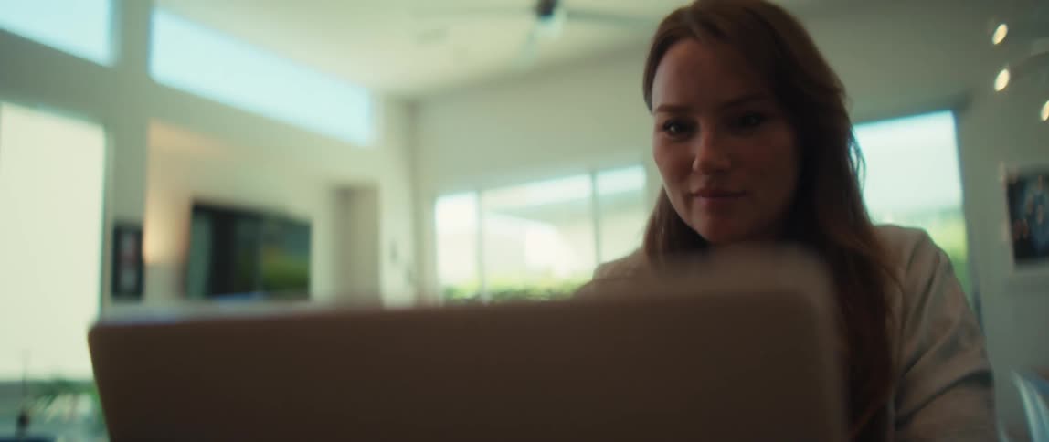 A young woman with auburn hair focuses intently on her laptop while working from her modern, sunlit home