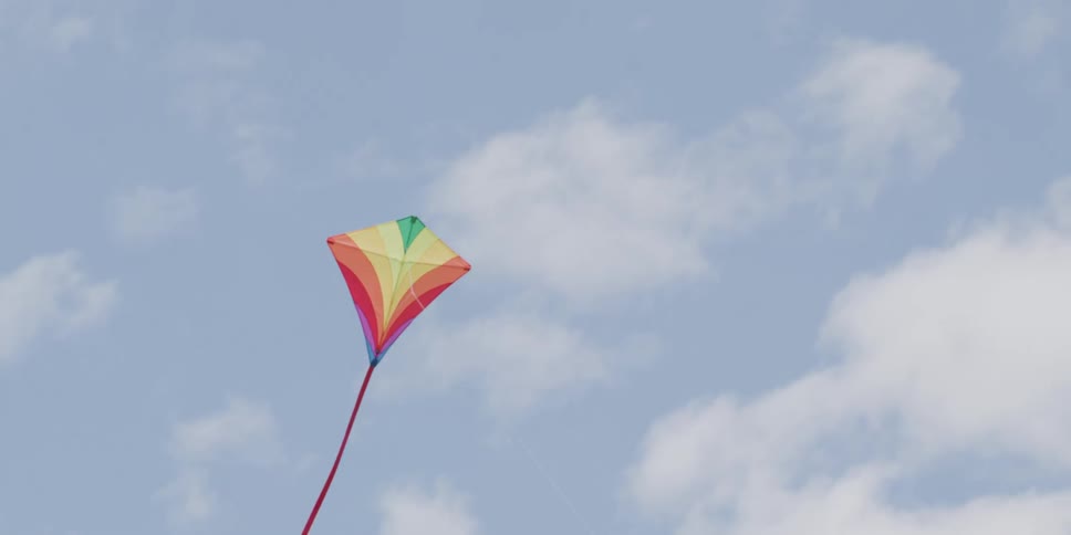 A colorful rainbow kite floats gently in the wind against a partly cloudy blue sky