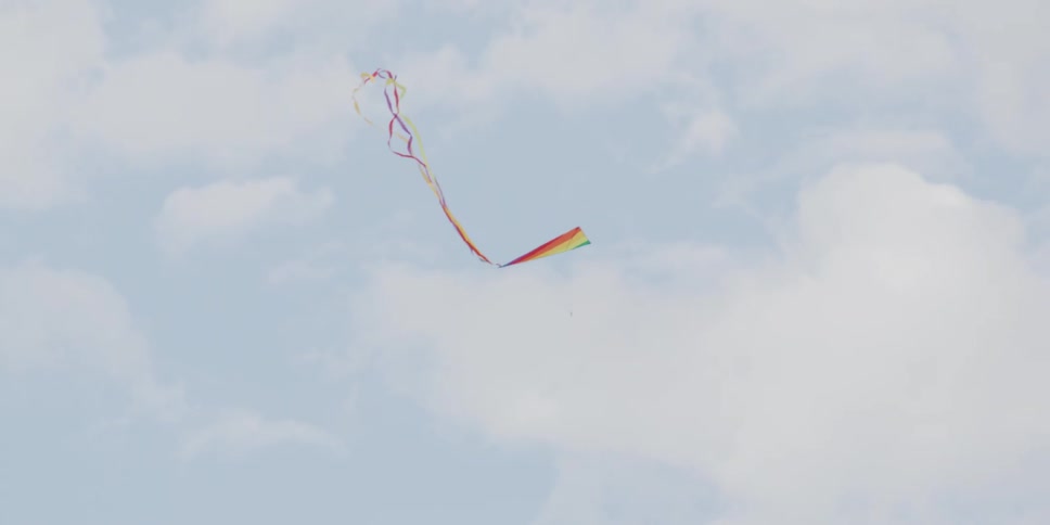 A colorful rainbow kite with a long flowing tail flies gracefully in slow motion against a bright and cloudy sky