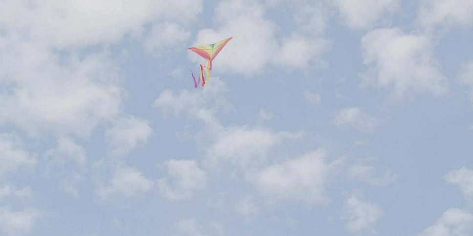 A colorful rainbow kite with long ribbons floats gracefully through a blue sky dotted with white clouds