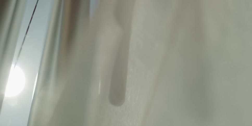 A tranquil, slow-motion close-up shows sunlight backlighting wind chimes and a sheer white curtain blowing in a gentle breeze