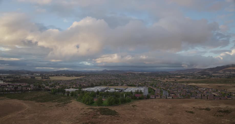 A wide aerial view shows a sprawling suburban community and school under a vast and cloudy sky