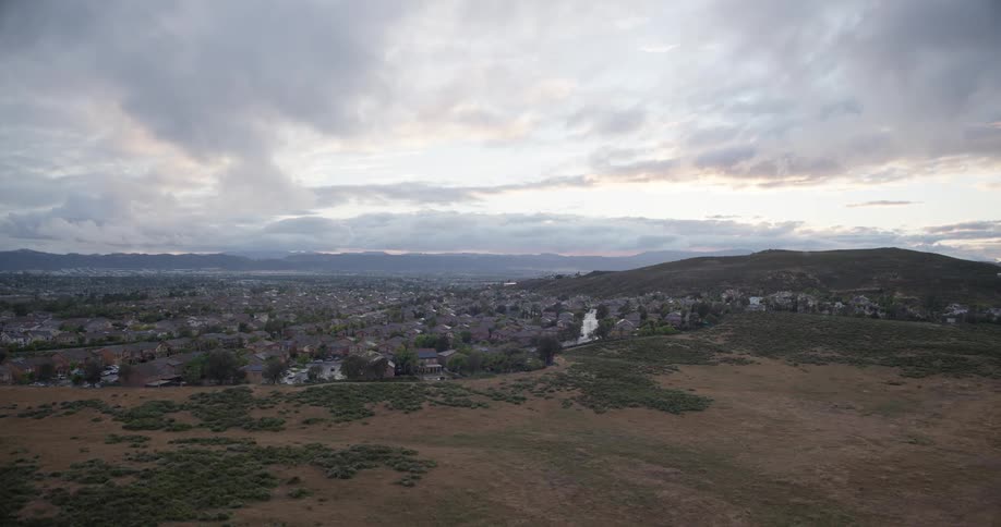 A slow aerial view tracks over a grassy field towards a suburban community nestled at the foot of rolling hills under a cloudy sky at sunset