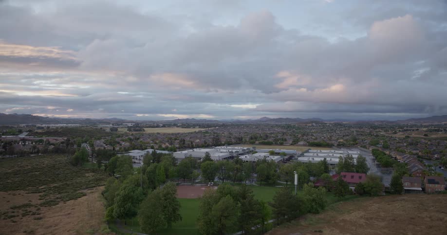 A slow aerial dolly shot pushes in from a dry field towards a suburban community park and school under a cloudy sky at dusk