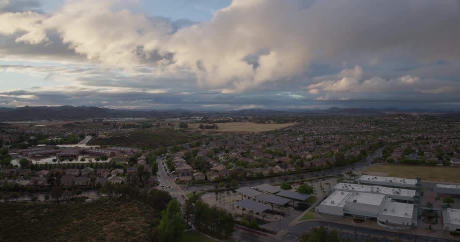 Expansive aerial pullback reveals a sprawling suburban community and schools under dramatic, sun-kissed morning clouds