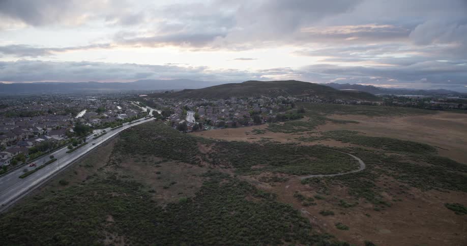 A serene aerial shot captures a suburban community nestled beside a highway and vast, undeveloped hills under a cloudy dawn sky
