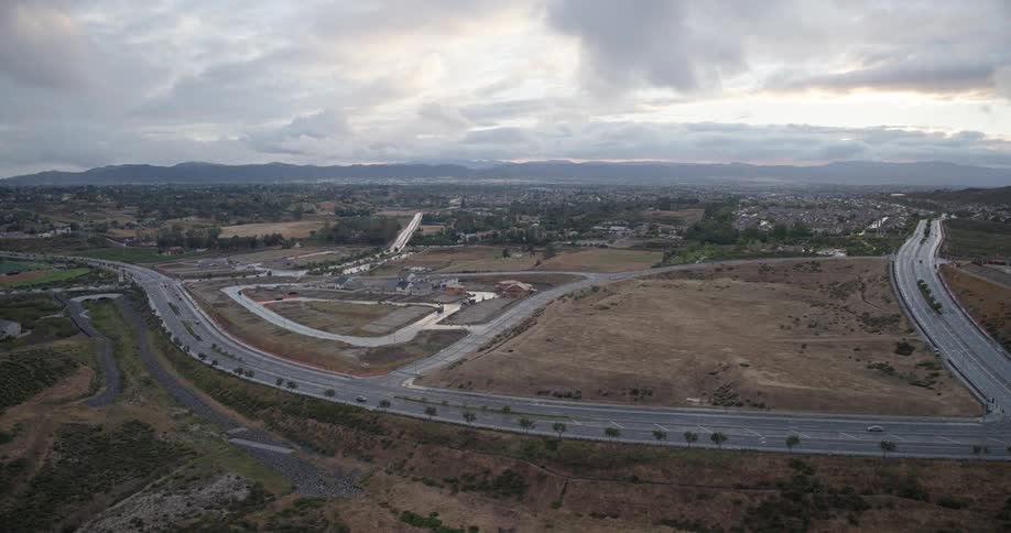 A slow aerial forward shot reveals a sprawling suburban landscape with new homes under construction and cars driving on winding roads under a cloudy sky