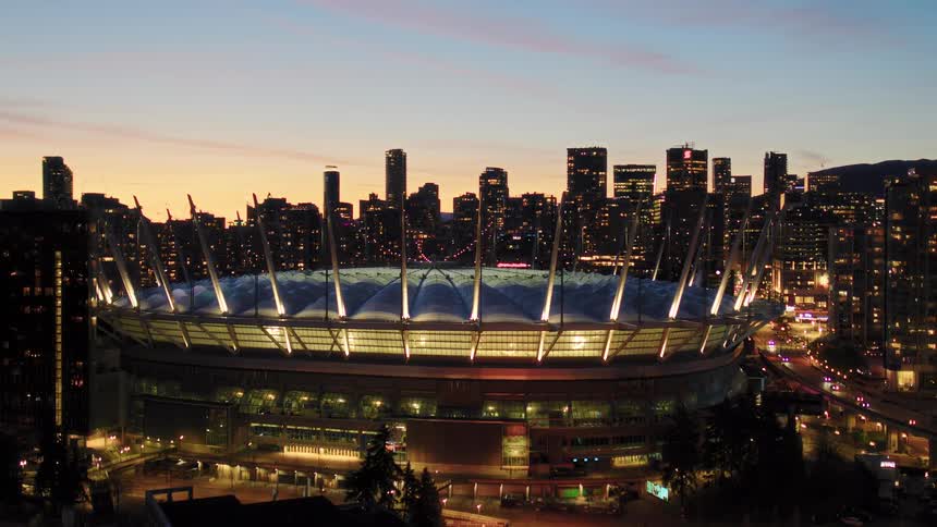 A stunning aerial view captures the iconic BC Place stadium brightly lit against the downtown Vancouver skyline at sunset