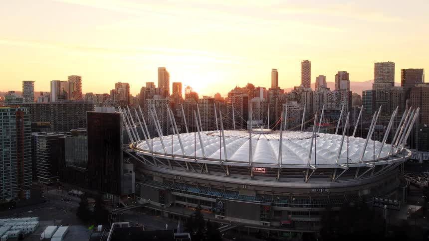 A cinematic aerial shot tilts up from the water's reflection to reveal the iconic BC Place stadium and the Vancouver city skyline during a stunning golden sunset
