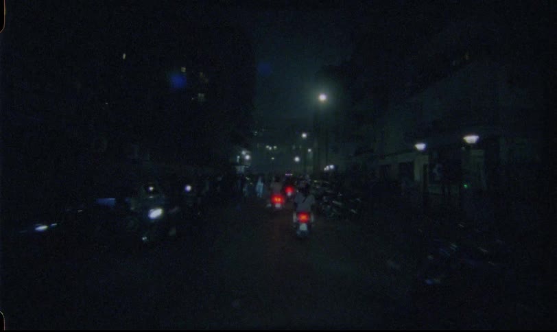 A grainy, vintage film shot captures a slow POV drive through a dimly lit urban street at night