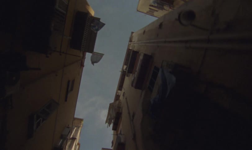 A low-angle tracking shot moves backward down a narrow alley, looking up at the sky between tall, weathered apartment buildings