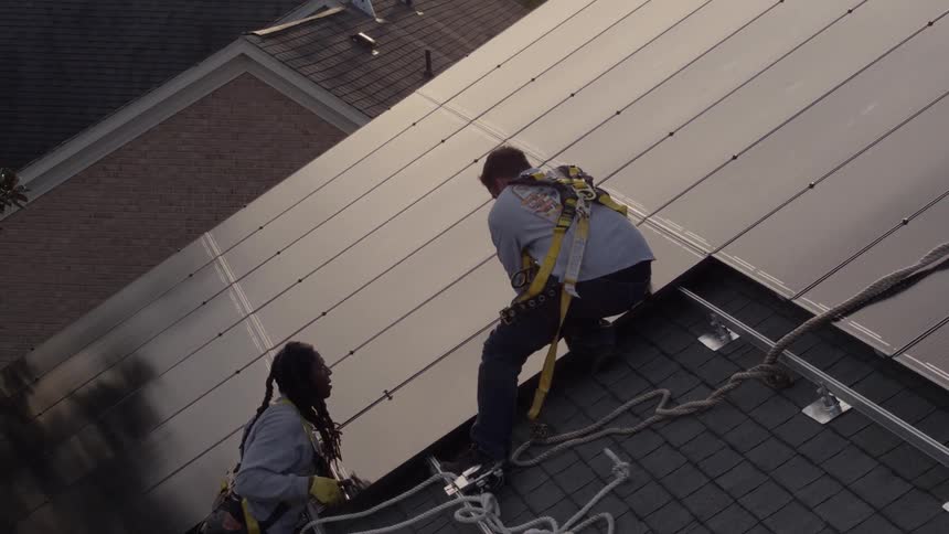 Two men wearing harnesses are installing solar panels on a house