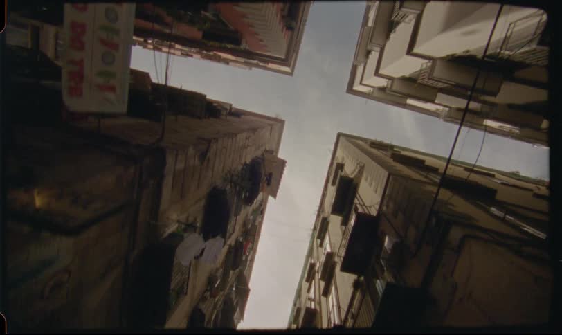 A low-angle tracking shot moves through a narrow alley in an old European city, looking up at the sky between tall apartment buildings