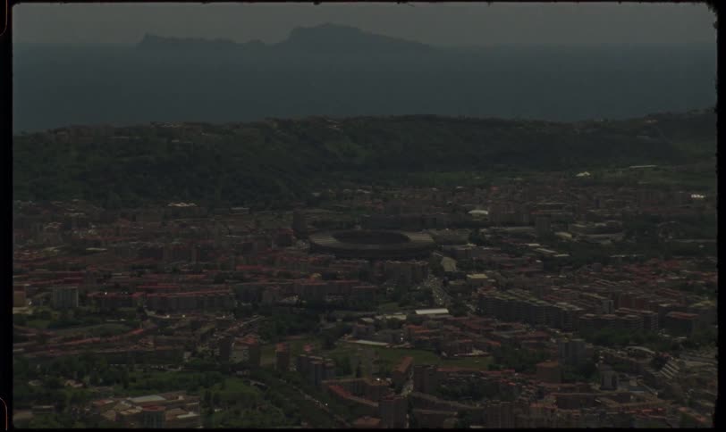 A vintage 1970s aerial view zooms out from a large stadium to reveal the sprawling coastal city of Naples, Italy, with the island of Capri in the distance