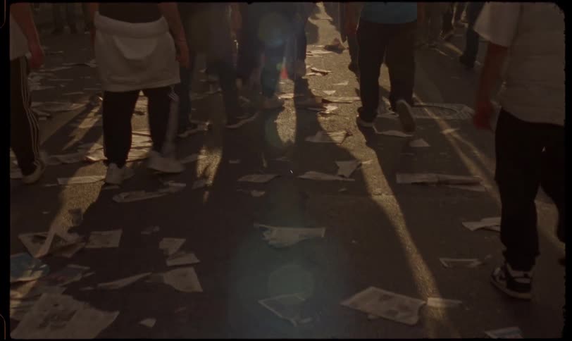 A crowd of people walk away in slow motion over a street littered with newspapers as long shadows are cast by the setting sun