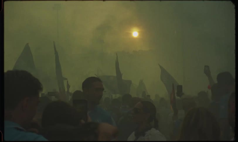 A vintage-style slow-motion shot captures a crowd of fans waving flags and recording on their phones amidst thick smoke at sunset