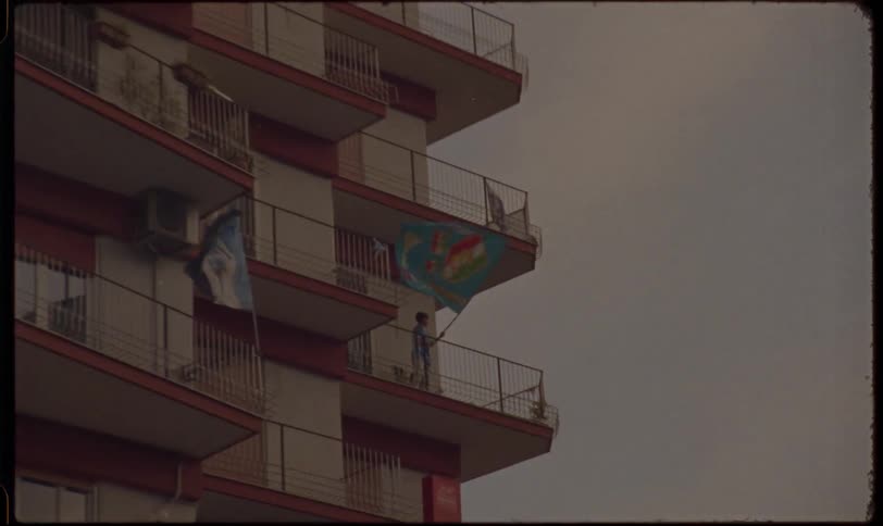 Vintage film captures a fan on a city apartment balcony waving a team flag in celebration against an overcast sky