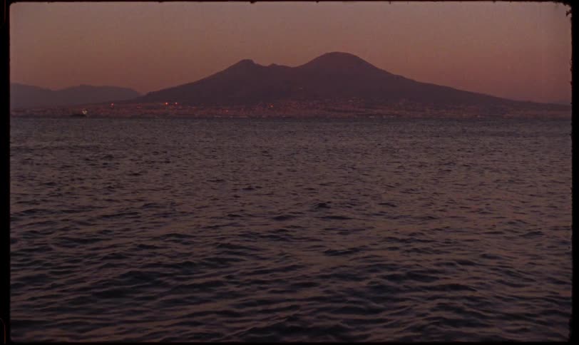 A vintage-style shot slowly zooms out to reveal Mount Vesuvius overlooking the Bay of Naples and the glowing city lights at dusk