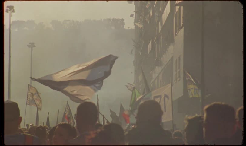 Vintage footage captures a massive crowd of passionate soccer fans waving flags through thick smoke in a golden hour haze