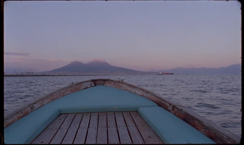 Point-of-view from a small wooden boat traveling across the calm bay of Naples toward Mount Vesuvius at dusk