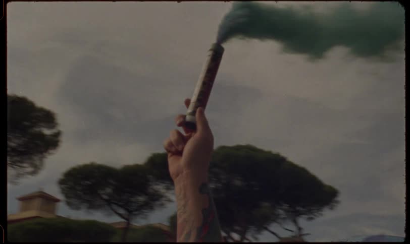 A vintage-style shot captures a tattooed hand holding a green smoke flare high in the air during a street celebration or protest