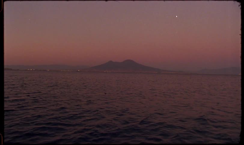 A vintage, grainy shot from the sea shows the silhouette of Mount Vesuvius against a beautiful pink and purple sunset sky