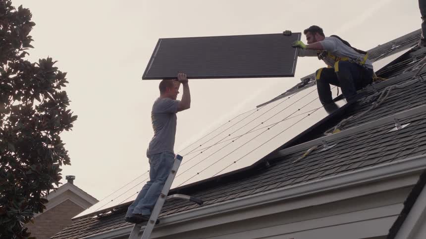 A man on a ladder hands a solar panel to a man on the roof
