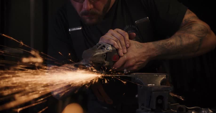 A tattooed metalworker uses an angle grinder in a dimly lit workshop, creating a shower of bright orange sparks