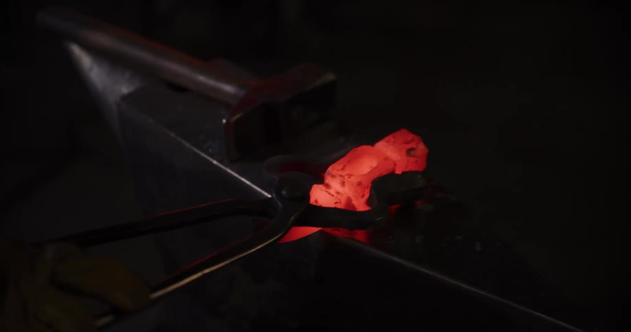 A blacksmith brushes and hammers a piece of glowing hot metal on an anvil in a dark workshop