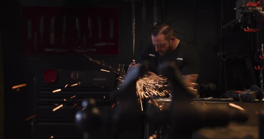 A craftsman in a dark workshop uses a power grinder on a metal object, sending a spectacular shower of sparks flying in slow motion