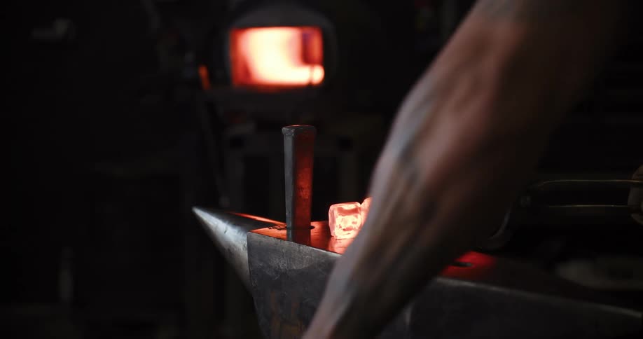 A blacksmith pulls a piece of glowing hot metal from a forge and hammers it into shape on an anvil in a dark workshop
