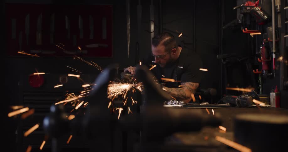 A tattooed craftsman works in his shop, using an angle grinder that sends a shower of sparks flying in slow motion