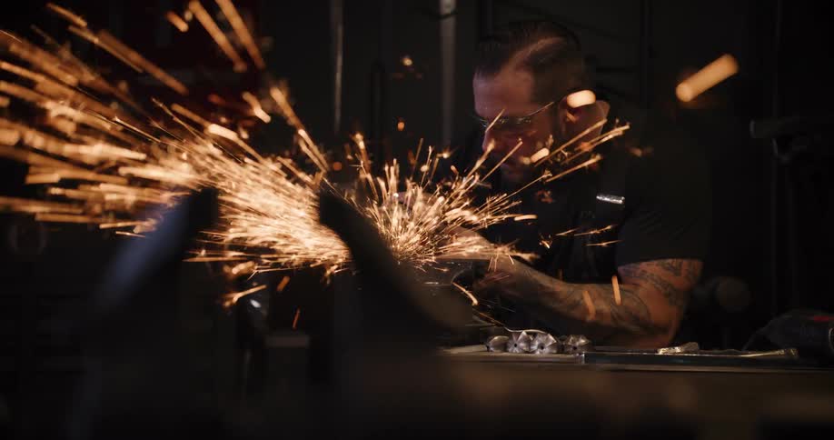 A craftsman wearing safety glasses uses an angle grinder on a piece of metal, sending a brilliant shower of sparks flying in slow motion through his workshop