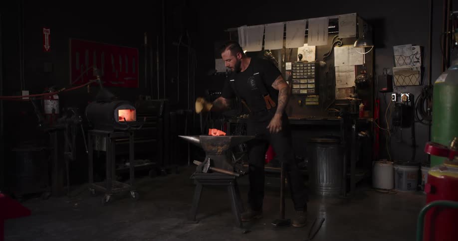 A skilled blacksmith with tattoos removes a red-hot metal billet from the forge and begins shaping it with a hammer on an anvil