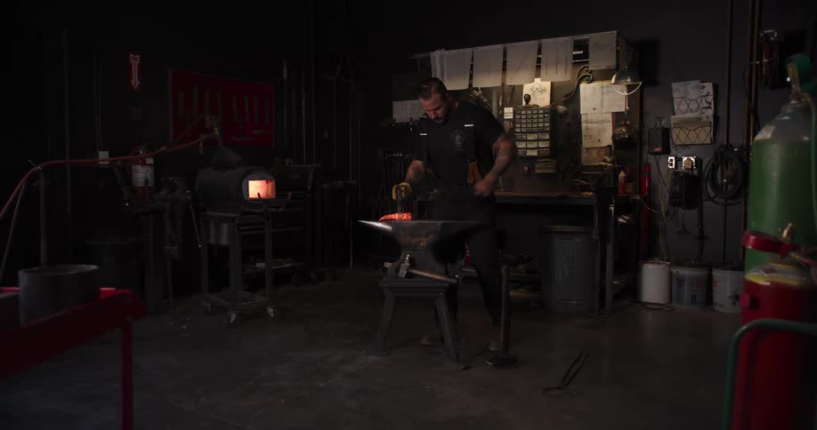 A blacksmith pounds a piece of glowing hot metal with a hammer on an anvil in his workshop