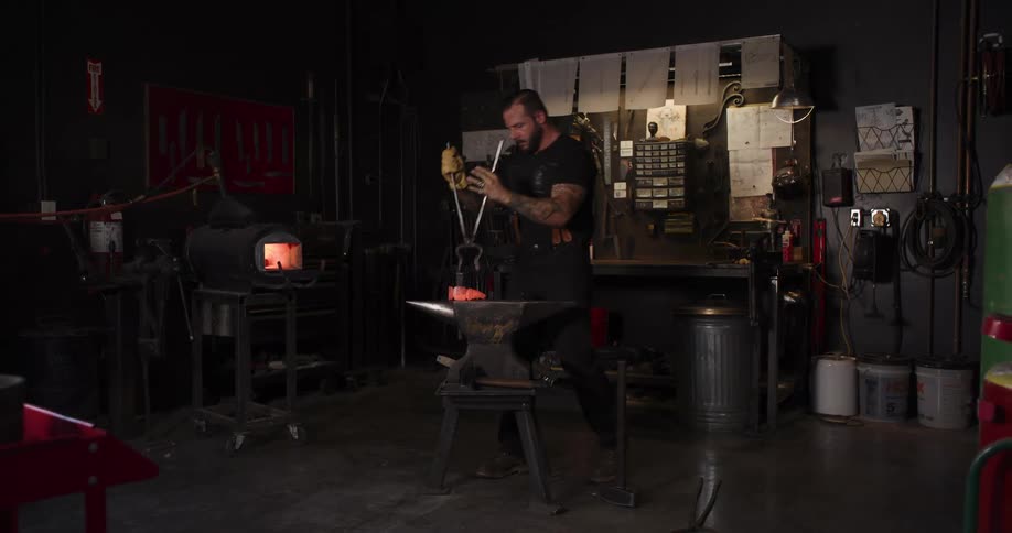 A strong, tattooed blacksmith hammers a piece of glowing hot metal on an anvil in a dark industrial workshop