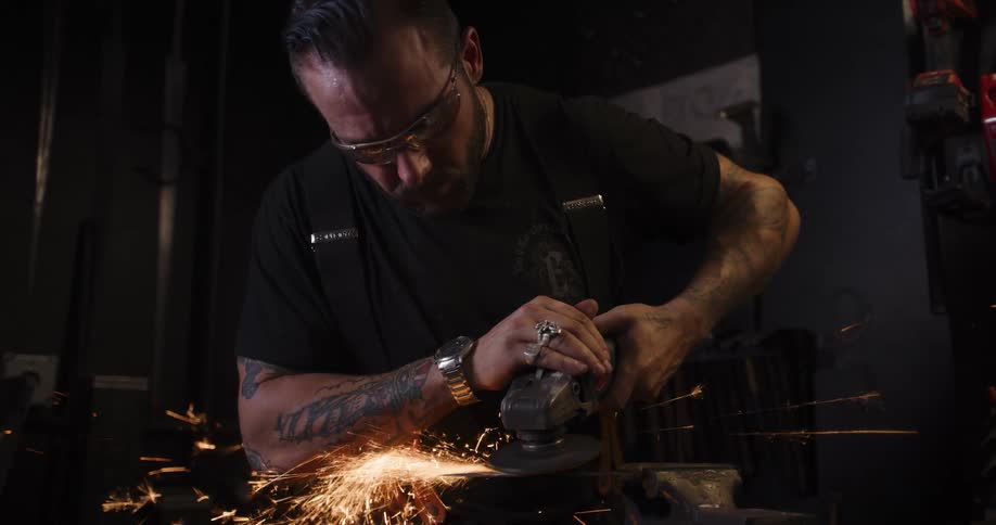 A tattooed metalworker in safety glasses uses an angle grinder on a piece of metal, creating a brilliant shower of sparks in slow motion