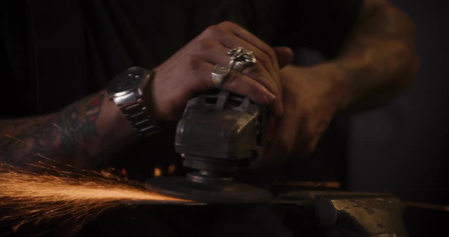 Tattooed craftsman grinds a metal blade with an angle grinder, creating a cascade of bright sparks in slow motion