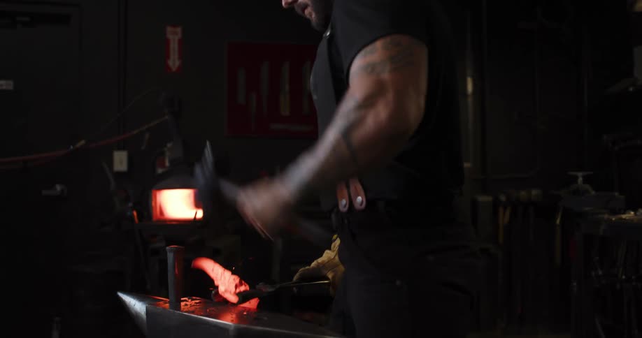 A strong, tattooed blacksmith hammers a glowing piece of metal on an anvil to forge a blade in his workshop