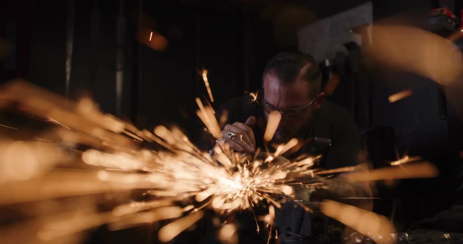 Mesmerizing cinemagraph of a tattooed craftsman grinding metal as a shower of fiery sparks flies towards the camera in a dark workshop